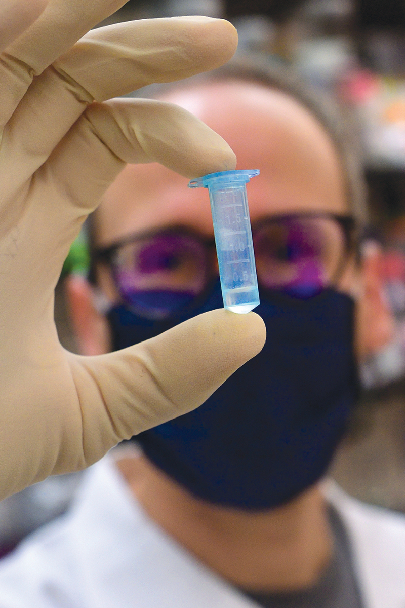 A doctor, with masked face blurred in the background, holds up a small, blue, graduated test tube in the foreground.