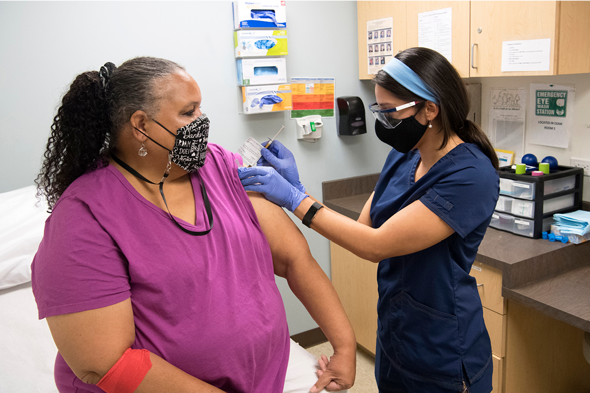 A woman wearing a purple shirt and a mask sits on an examination table and receives a shot from a female healthcare worker in blue scrubs, gloves, and headband.