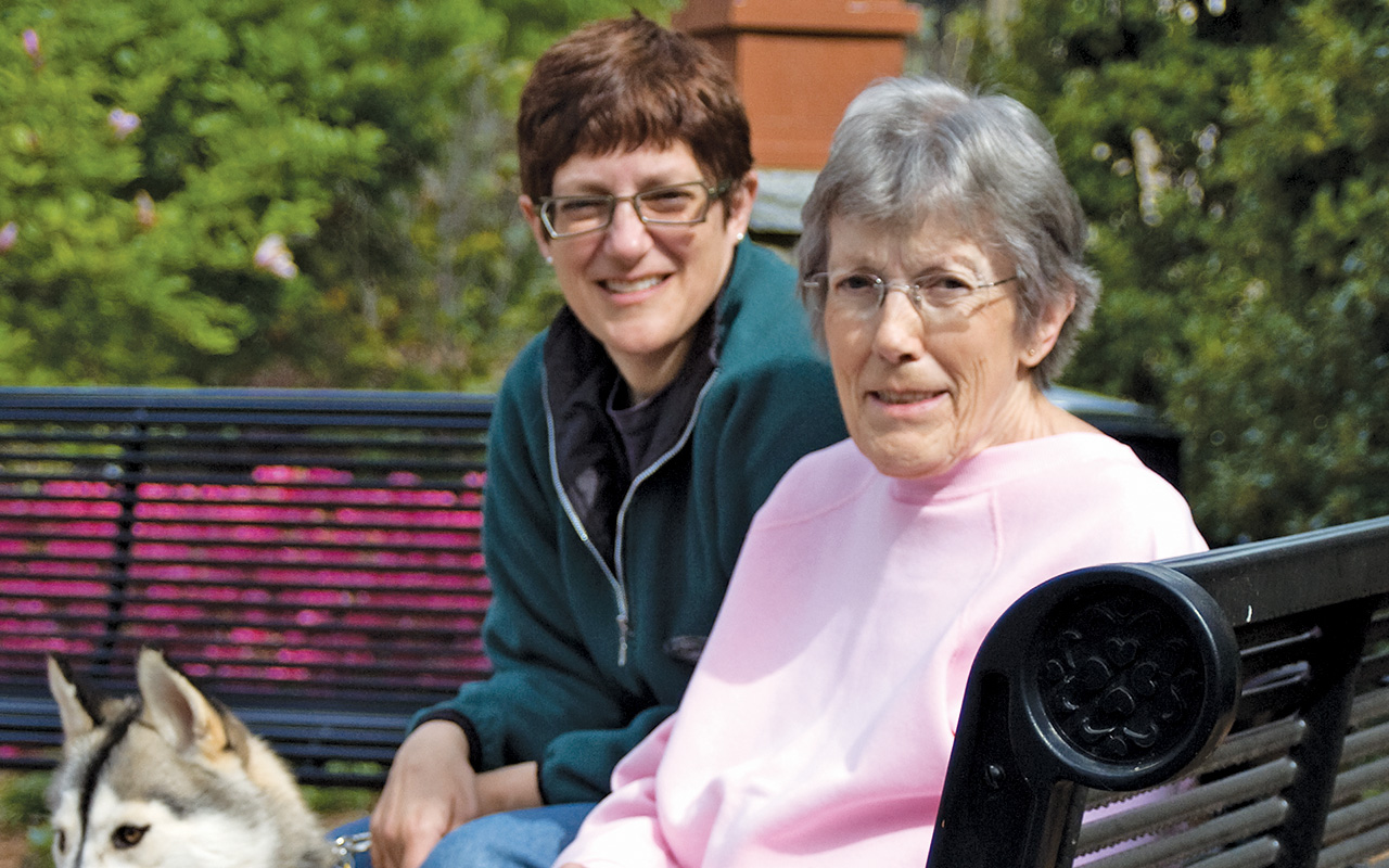 Pat and Susan Carini sit on a black, ornate metal bench with Susan's husky Sable beside them. It is Spring and azaleas bloom in the background.