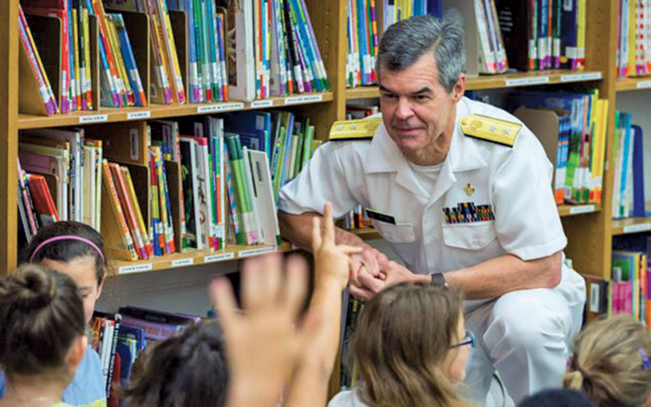 Stephen Redd in a white military style uniform kneels next to bookshelves. A group of children is in the foreground with their arms raised.