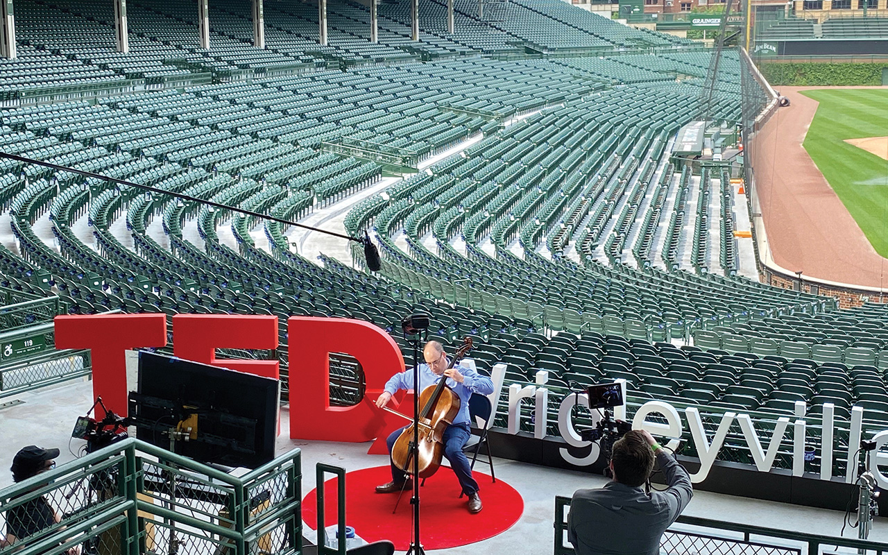 A man plays cello above empty seats at Wrigley Field. TEDxWrigleyville is spelled out in large cutout letters behind him.