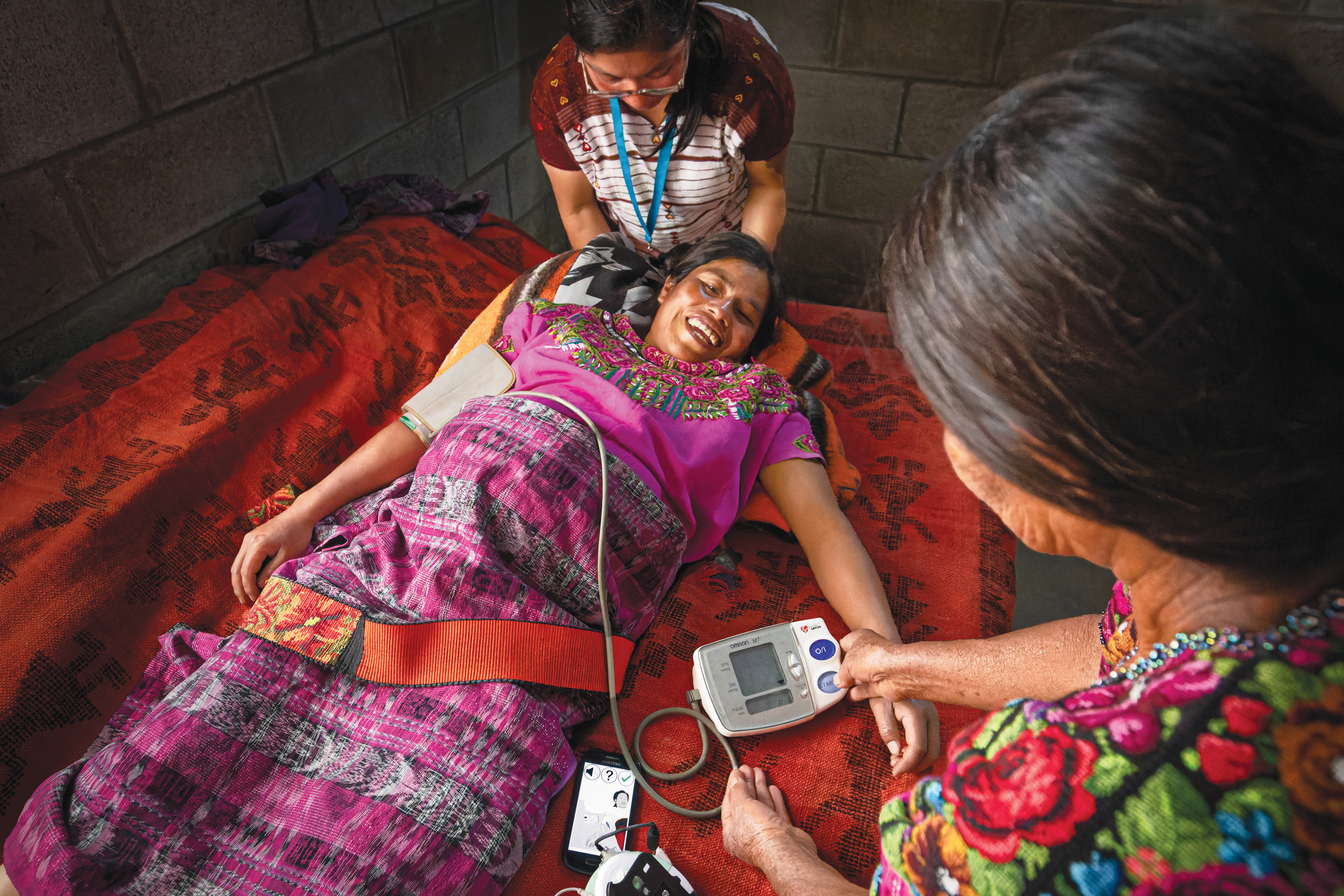 A colorfully dressed woman lies on a blanket while being attended to by a woman using a smartphone