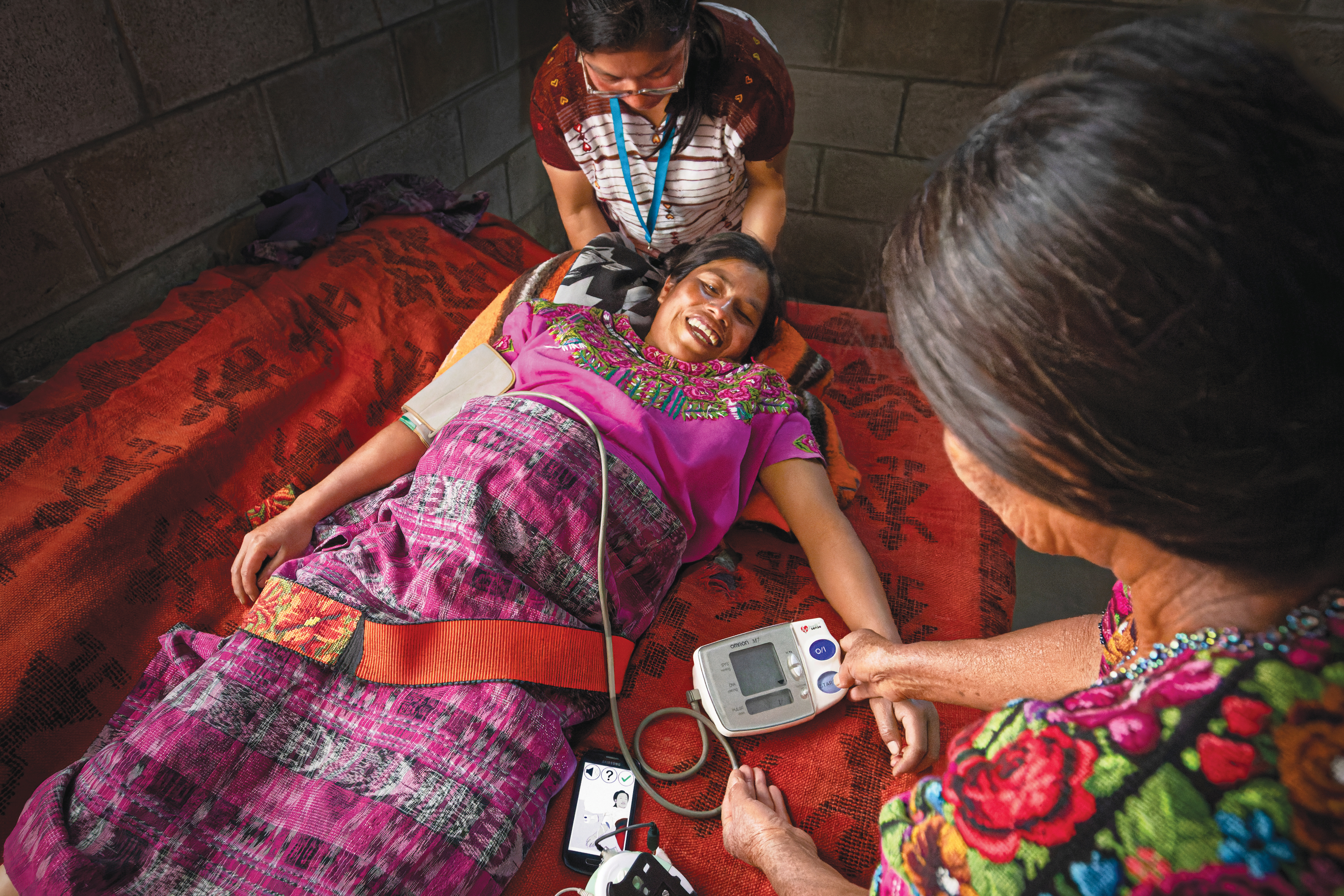  a pregnant, colorfully-dressed woman lies on a large piece of fabric receiving medical tests