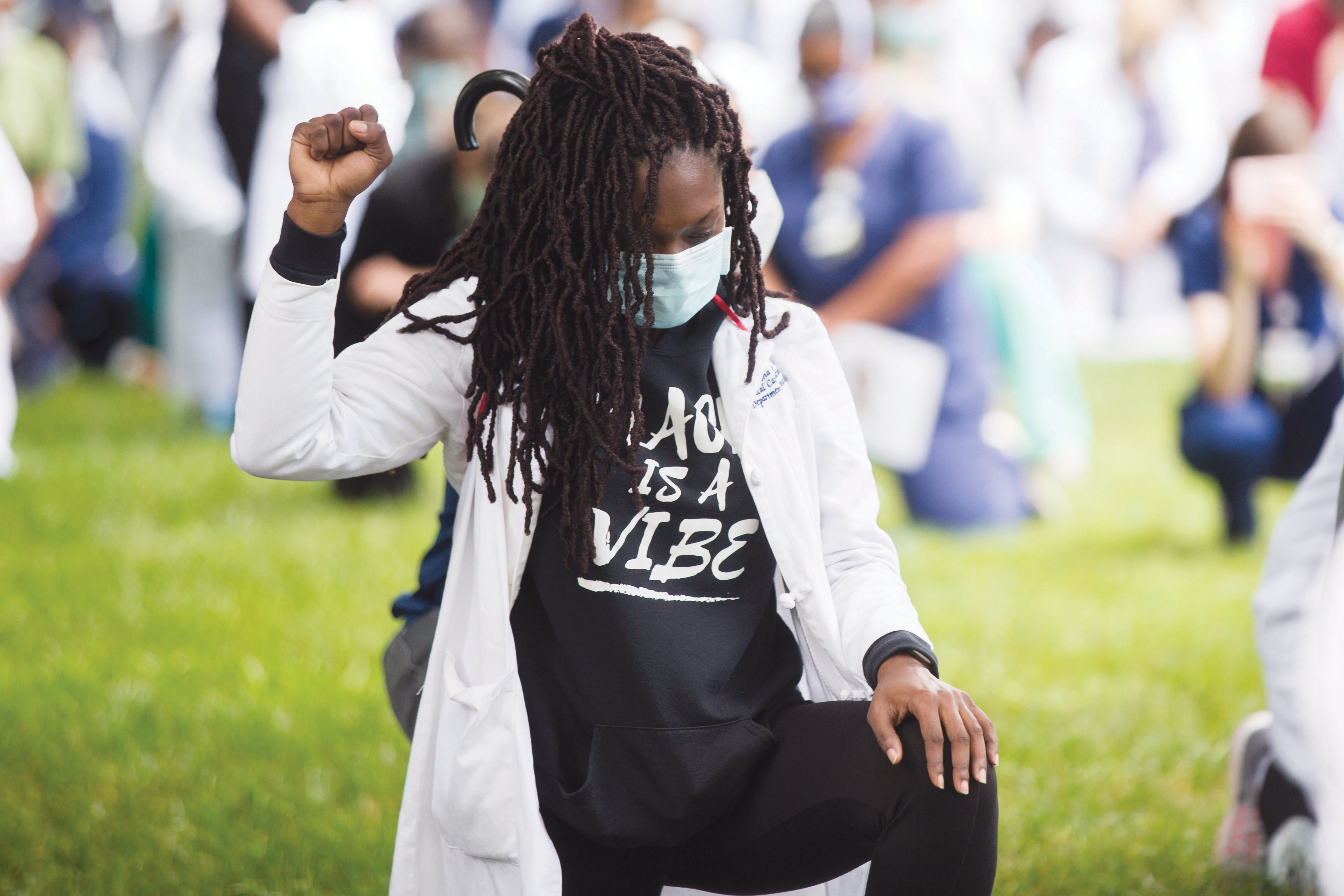 A masked student kneels on grass with her right hand raised